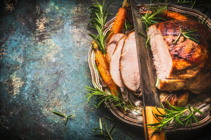 Roasted pork ham with kitchen knife and roast vegetables on dark rustic background, top view, border
