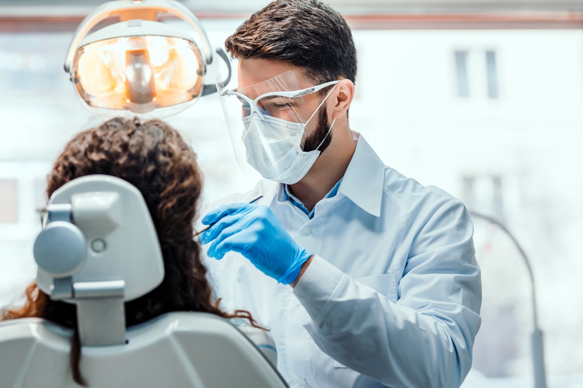 Male Dental Practitioner with brown hair, a mask and clear visor is looking at a female patient with brown curly hair who is sitting in a white dental chair.