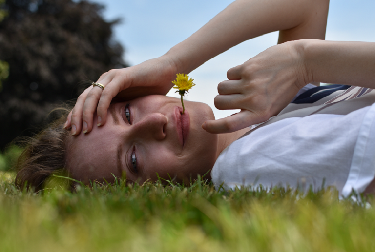 A woman lying on grass with a dandlion between her lips looking at the camera