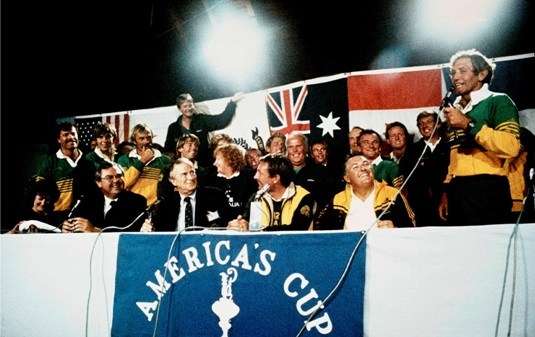 Group of Australians at a press conference desk, Australian flags in background, America