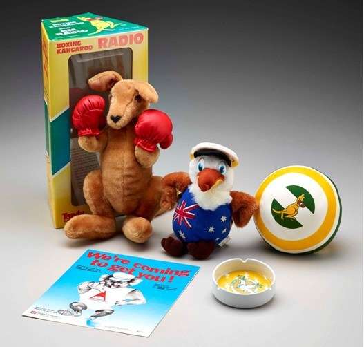 Photo of souvenirs: Plush boxing kangaroo toy, plush kookaburra toy, small ball with boxing kangaroo graphic, ashtray with yellow kookaburra graphic, poster with koala graphic and slogan