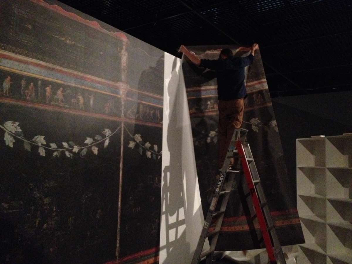 The graphic of the dining room fresco from the Villa Farnesina in Rome, 20–15BC being installed in the exhibition space where the body casts are on display. Fresco image courtesy the National Roman Museum – Palazzo Massimo alle Terme. Image: ANMM.