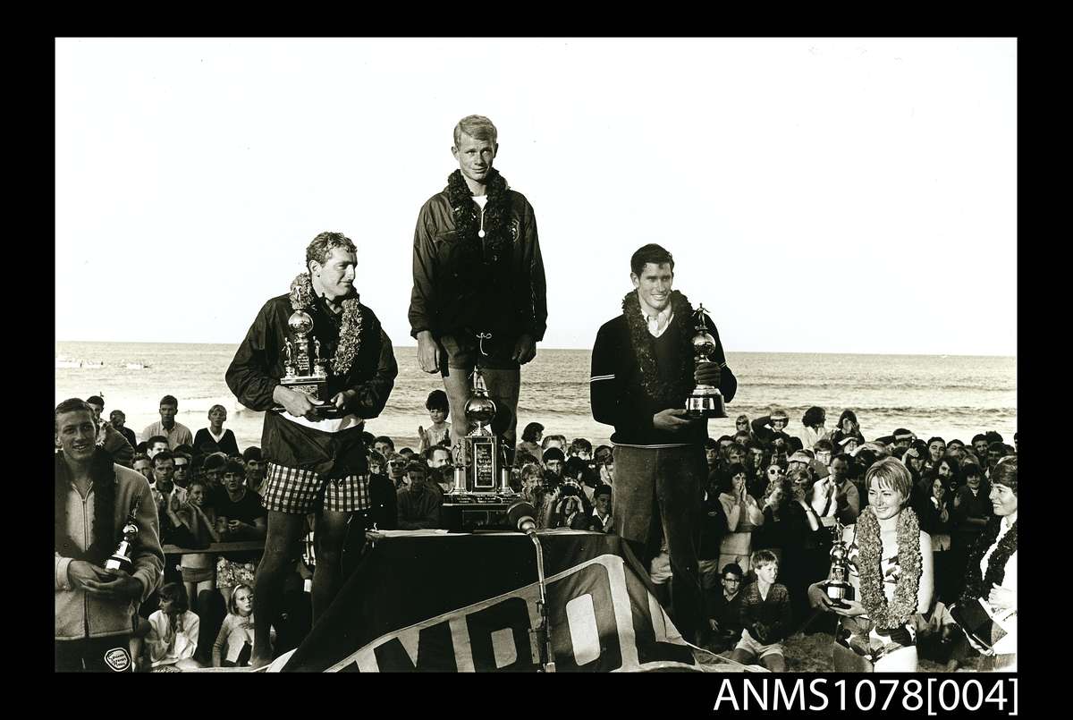 Jack Eden, Presentation of the trophies in the first world open surfboard championships Manly, 1964. Gelatin silver photograph, printed 1990s. Senior men’s championship winners on the podium: 1 Midget Farrelly, Australia (centre), 2 Mike Doyle, California (left), 3 Joey Cabell, Hawaii (right). The women’s championship winners are on the right: 1 Australian Phyllis O’Donnell (far right) and 2 Linda Benson California (right), with Nat Young, the runner-up in the junior men’s final, on the left. ANMM Collection ANMS1078[004], ANMM Collection Gift from Jack and Dawn Eden.