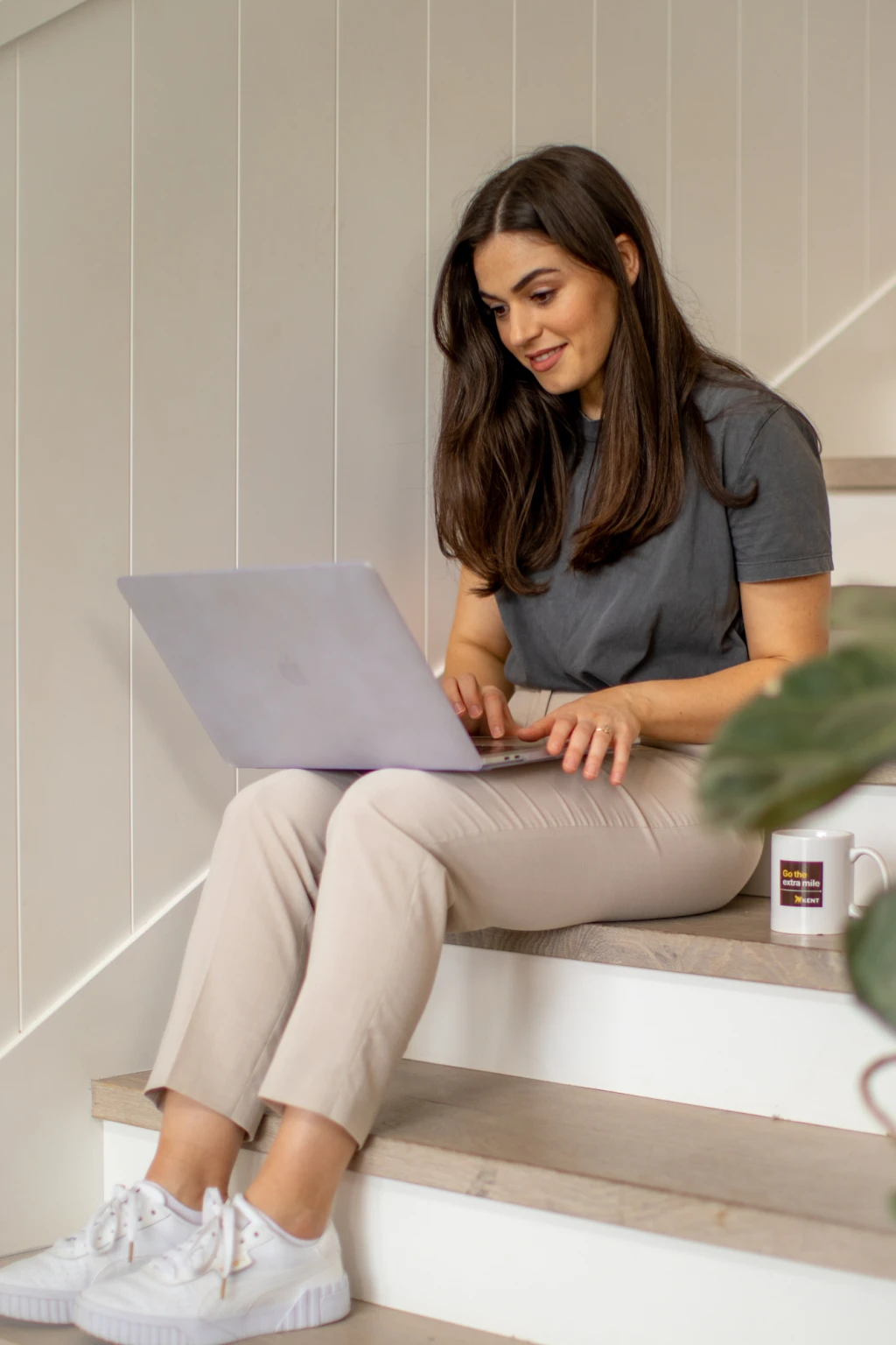 Woman sitting on stairs working on a laptop with a coffee mug beside her