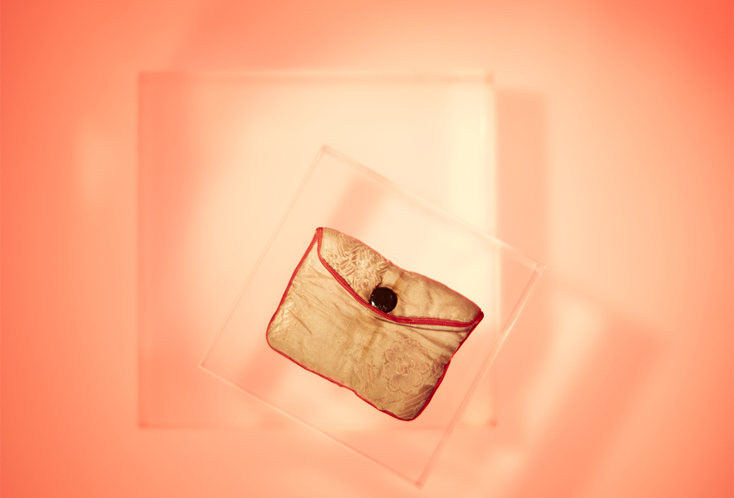 Pouch: Pink Silk, Vietnam pre 1978, Small floral pink silk pouch edged with red ribbon and closed with one metal button. Orange lit background, courtesy Immigration Museum, Melbourne