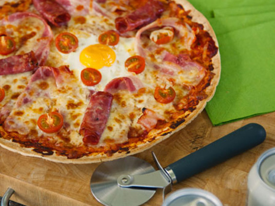 A breakfast pizza with a pizza cutter lying next to it on a wooden bench