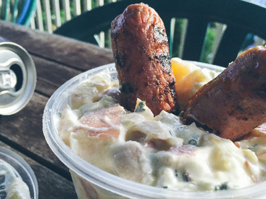 A small bowl of potato salad with two sausages stuck in it sitting on an outdoor table