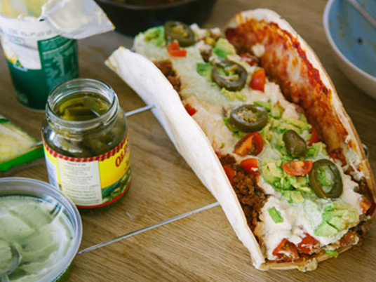 An italian taco with a jar of pickles next to it on a wooden bench