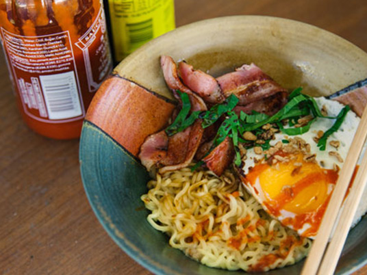 Noodles, egg and bacon in a bowl with a pair of chopsticks laying across the bowl