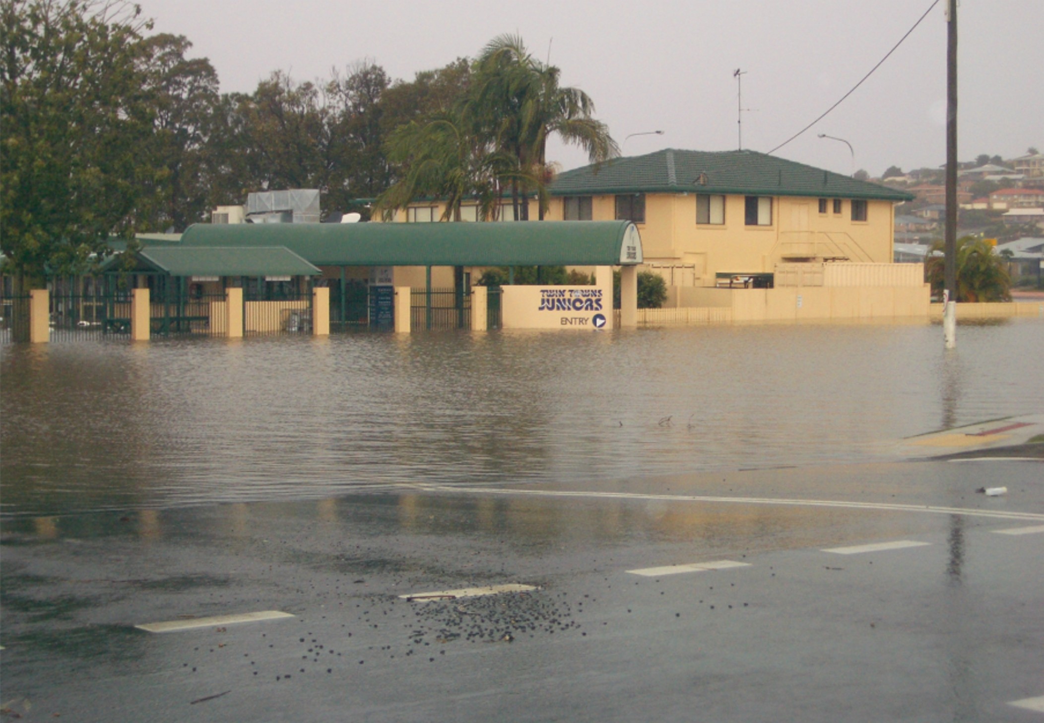 June 2005 flood: Twin Towns Juniors, Fraser Drive and Leisure Drive, Banora Point. The image shows a building partially submerged in floodwater. The water level is high, covering the road and reaching up to the entrance of the building. Trees and other structures are visible in the background.