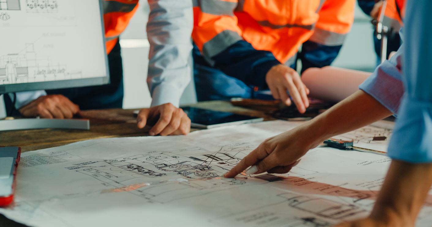 Workers in high-vis vests point at engineering plans on a table, symbolising planning discussions.
