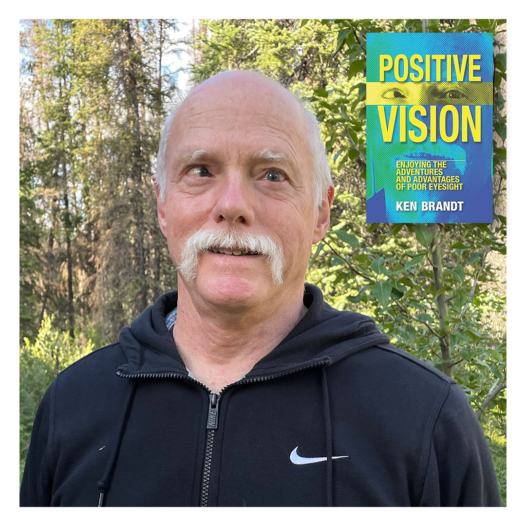 Photo of Ken Brandt, a bald man with a white moustache, standing outdoors in front of green trees, wearing a black Nike zip-up hoodie. In the top right corner is the cover of his book Positive Vision: Enjoying the Adventures and Advantages of Poor Eyesight by Ken Brandt. 