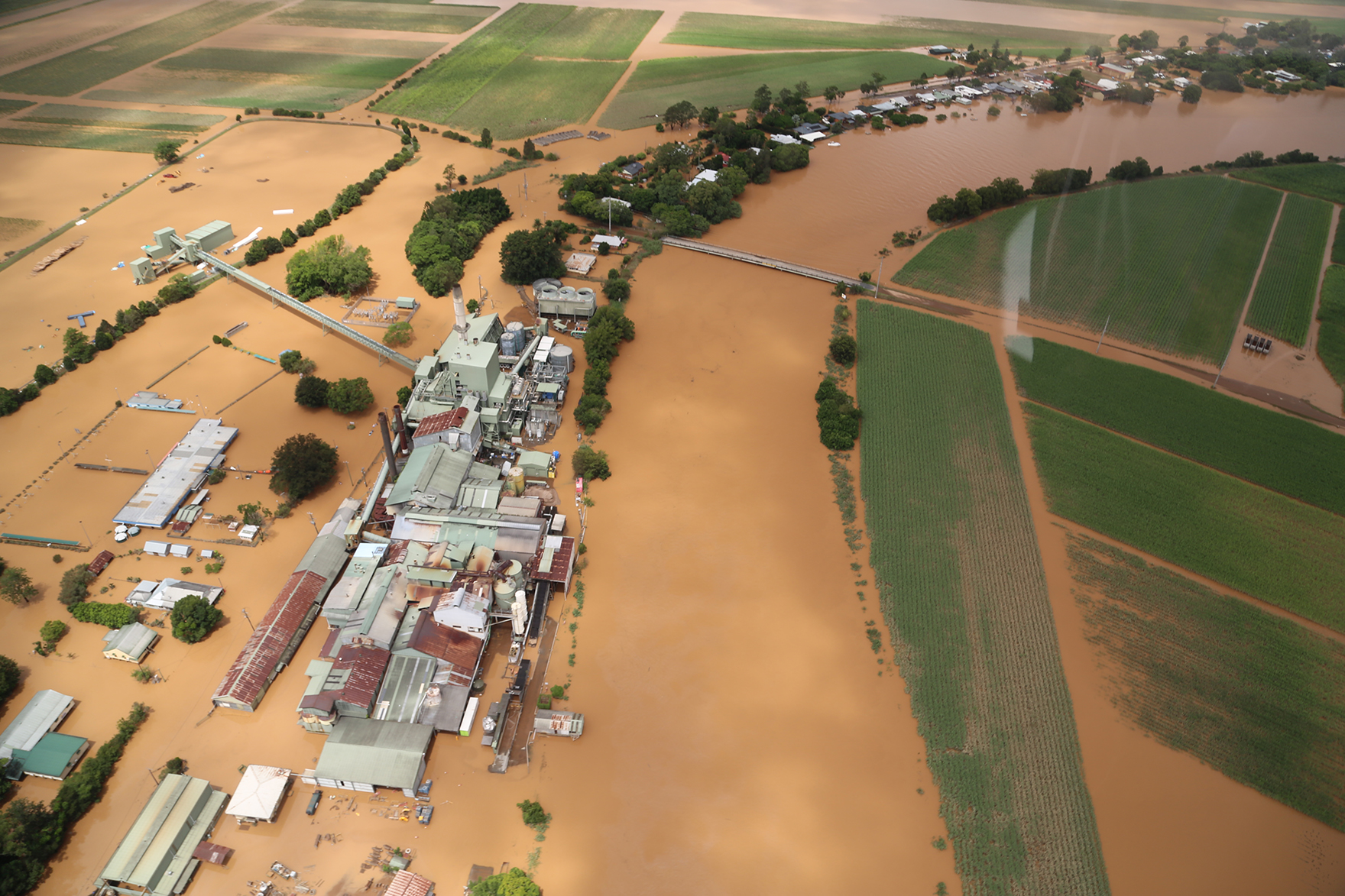 Aerial photograph showing extensive floodwaters inundating the Condong sugar mill and adjacent agricultural fields, with muddy water submerging roads and nearby properties.