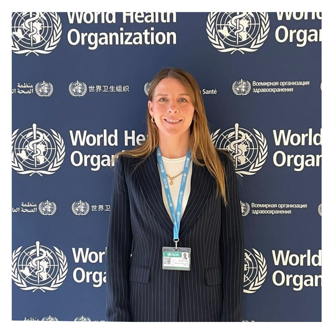 Dr Kelsey Chapman a young white woman with long blonde hair and wearing a navy pinstripe pants suit and lanyard stands in front of the WHO photowall at the conference.