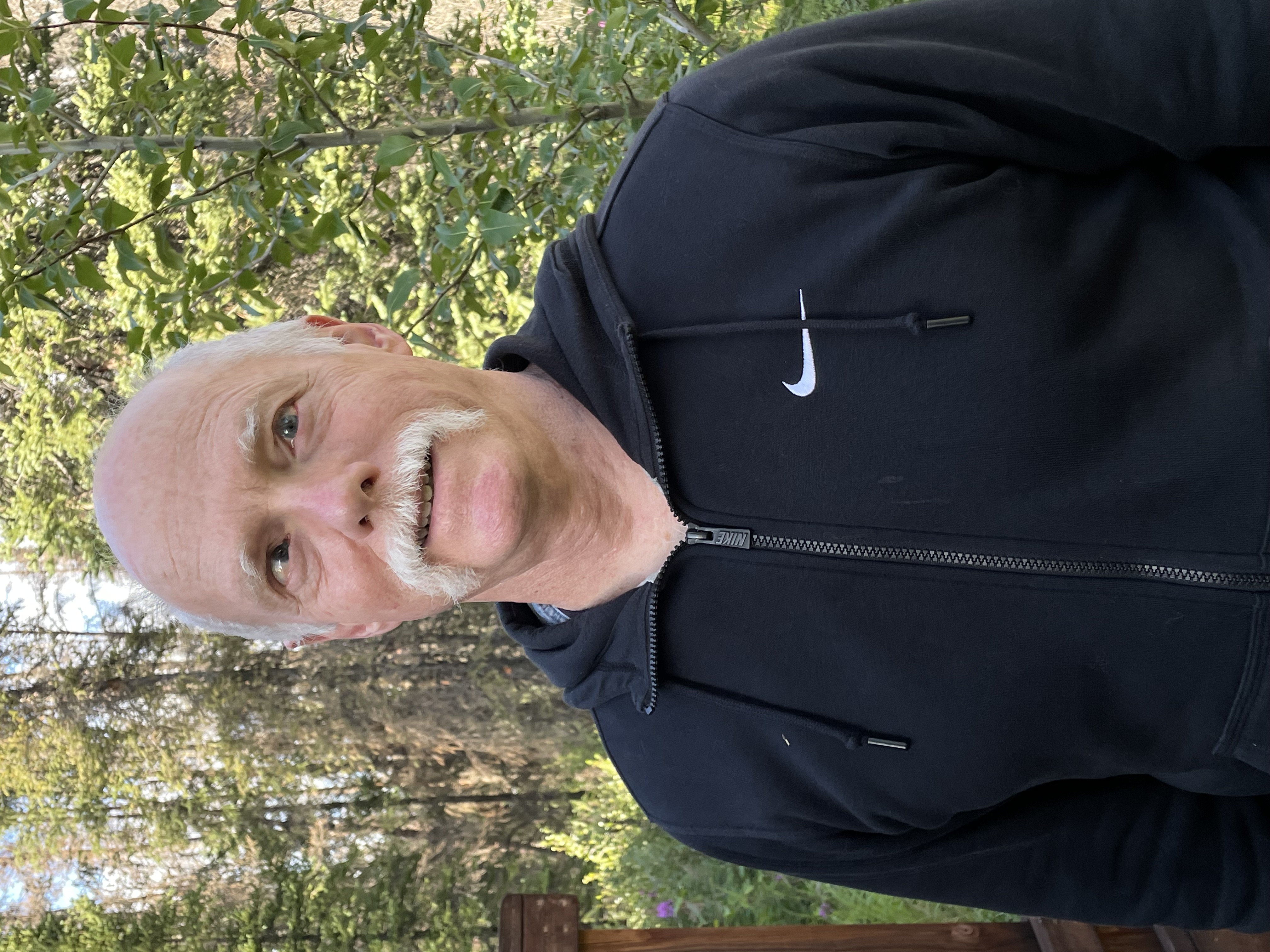 A man standing facing the camera wearing a black sports jacket. He is balding and has a moustache. Behind him is a forest with sunlight through the trees. 