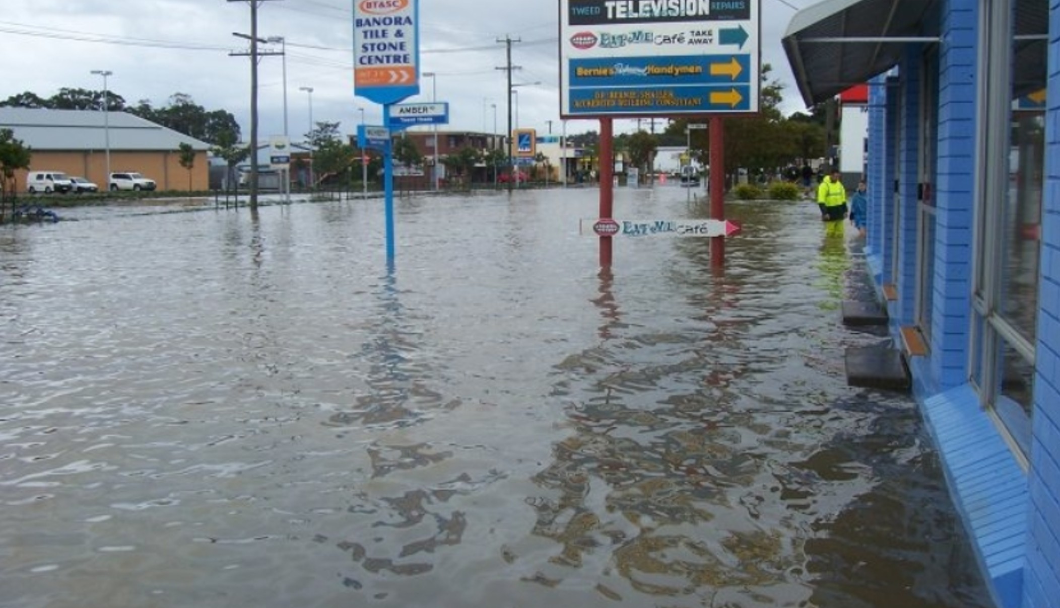 June 2005 flood: Machinery Drive, Tweed Heads South. The image shows a flooded street with water covering the entire road and reaching up to the buildings. Several signs are visible, including one for 'Tweed Television' and another for 'Banora Tile & Stone Centre'.