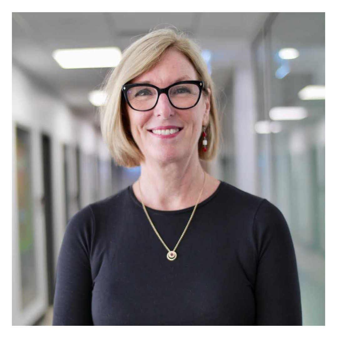 Dr Kathy Gibbs from Griffith University smiles at the camera while standing in a bright hallway. She has short blonde hair and wears black-framed glasses, a black top, a gold necklace, and red earrings.