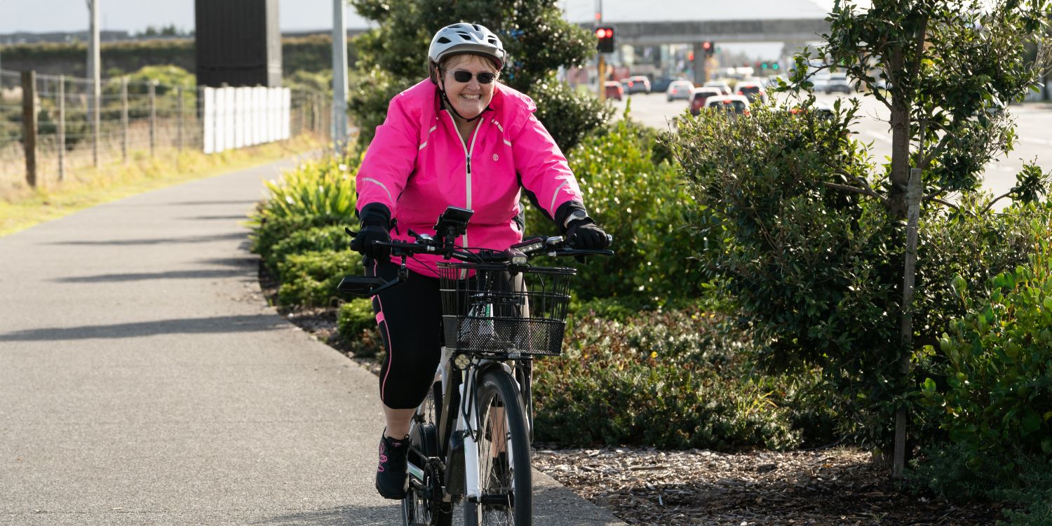 A woman biking along the cycle path in Kāpiti with cars and traffic lights in the background.