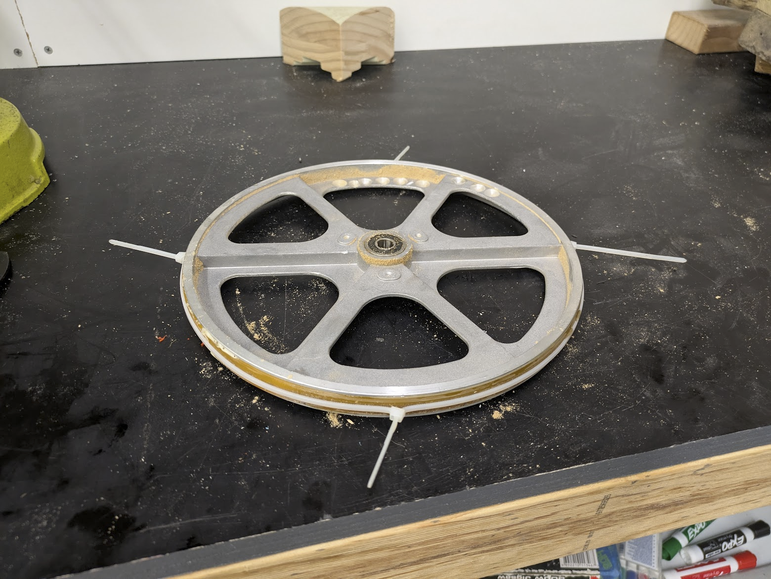 A photo of a spoked aluminium wheel sitting on a black workbench. There is a yellow-brown tyre running around the circumference, held on by some zip-ties. There is sawdust everywhere. The wheel is about 250mm in diameter and 20mm thick.