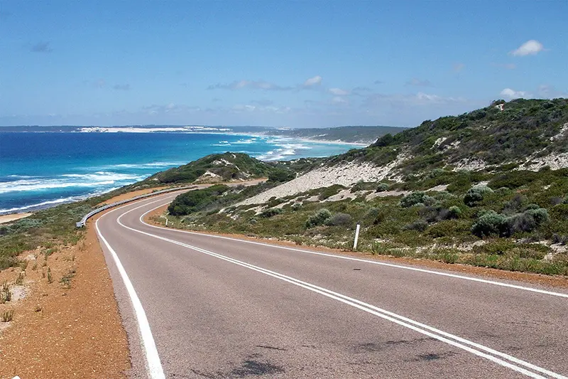 Coastal road winding along the shoreline with ocean waves on one side and green hills on the other under a clear blue sky