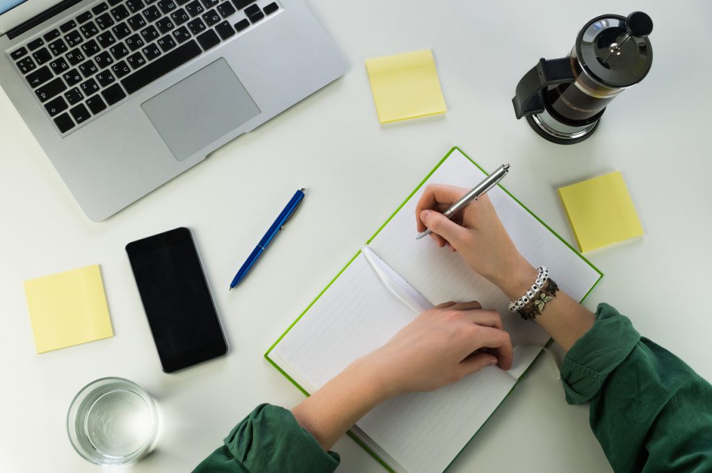 Writing in notepad sitting at desk with smartphone and laptop computer