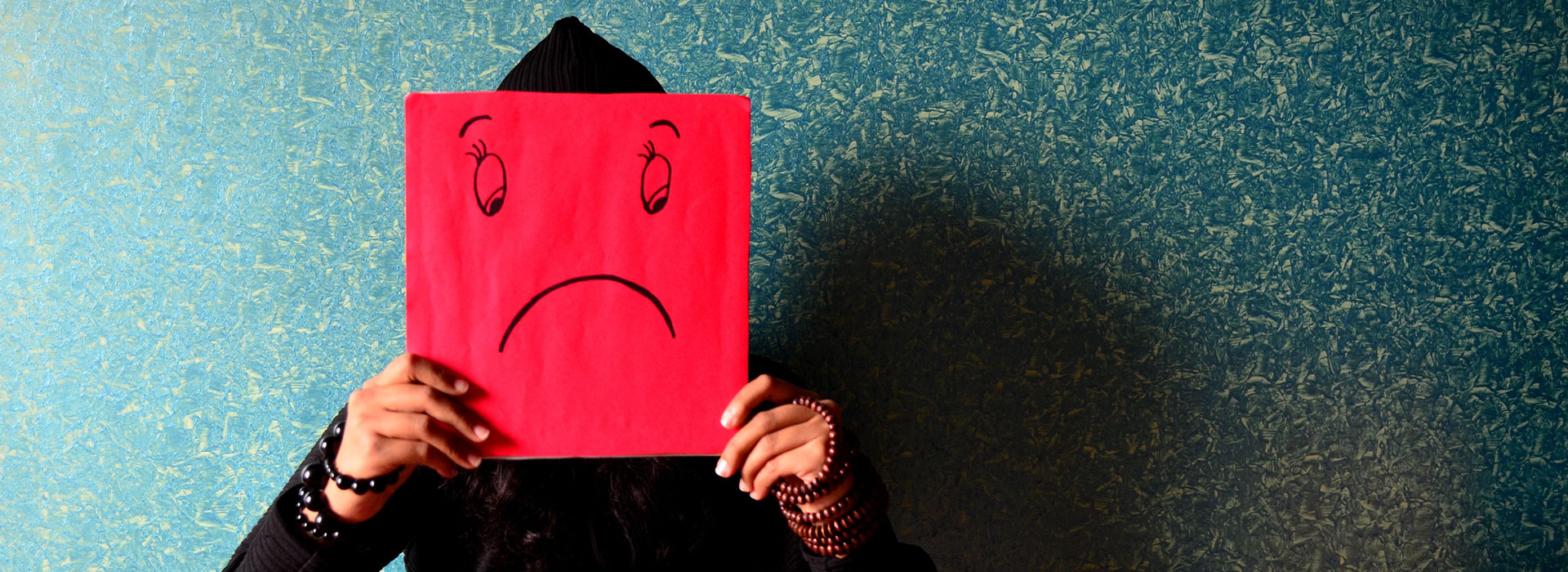 A person holding up a red square in front of their face with a sad face drawn on it.