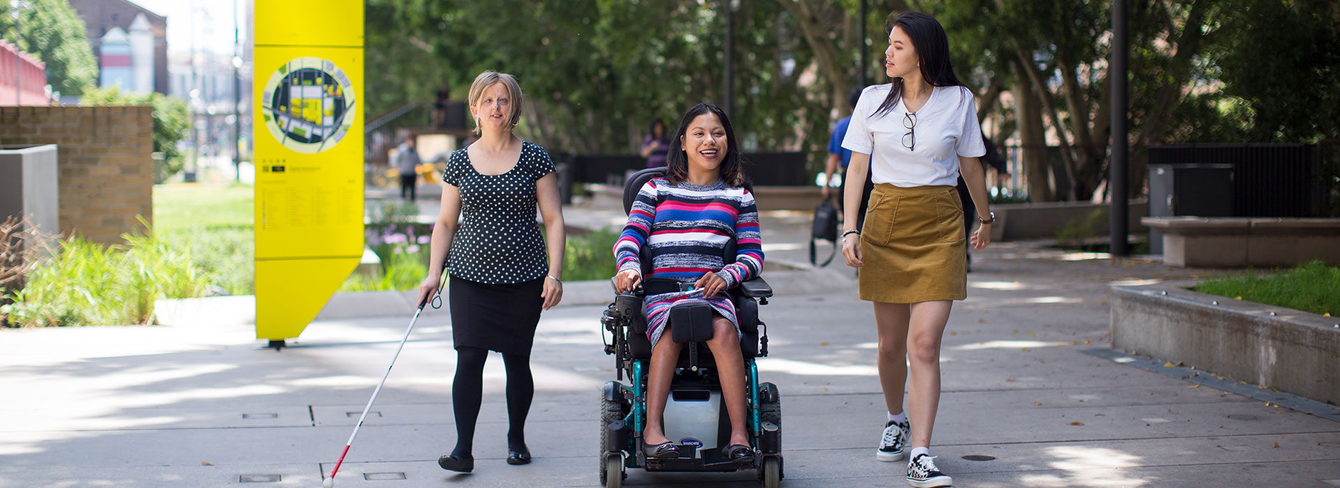 Three women walking alongside each other. One is vision impaired and is holding a stick, one is in an electronic wheelchair and the other is able bodied.