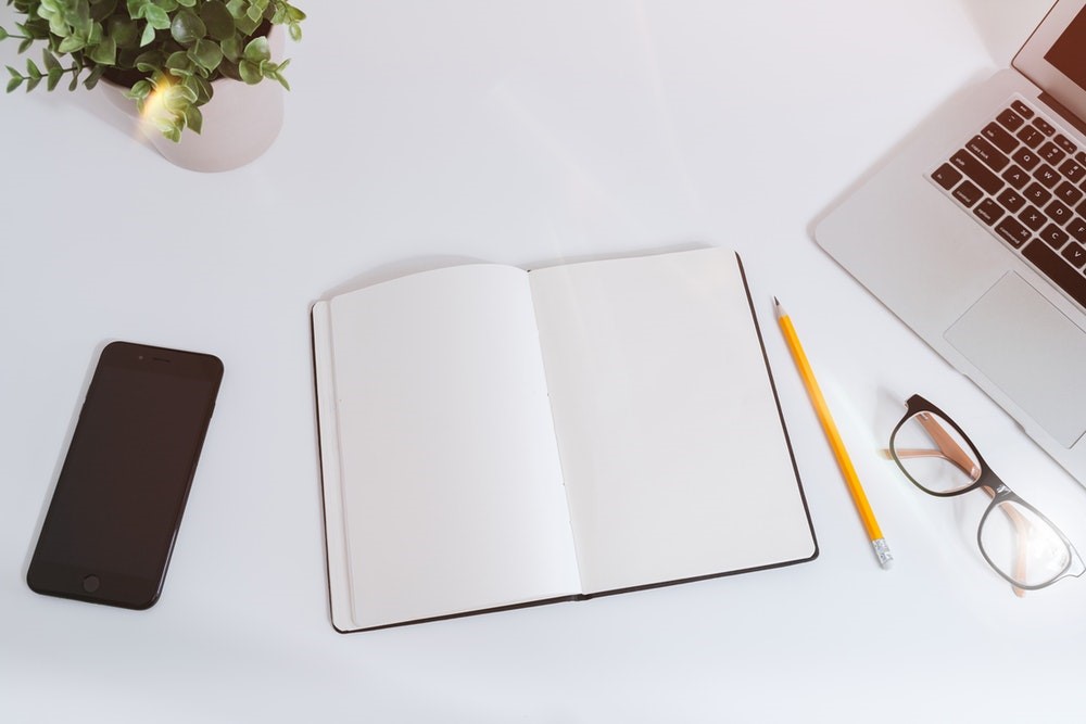 Aerial flatlay of black open notebook, phone, laptop, pencil, glasses and plant