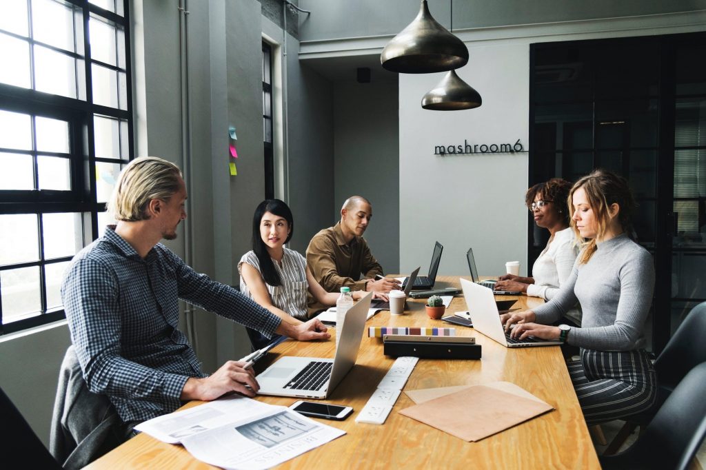 five people in their 30s sitting around a desk with laptops