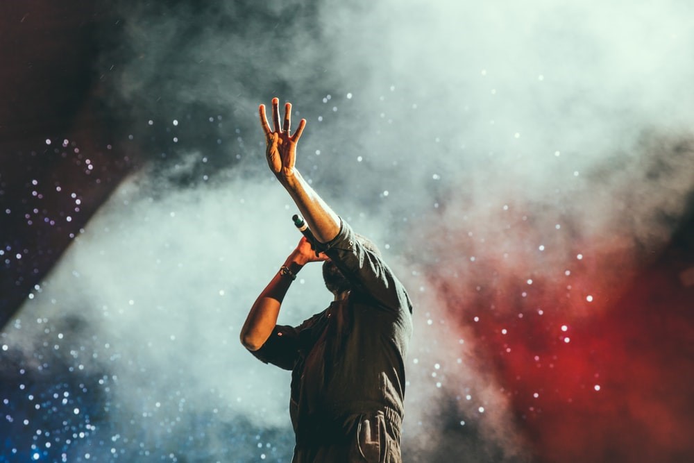 man with microphone, left hand in air obscuring face in front of smokey background